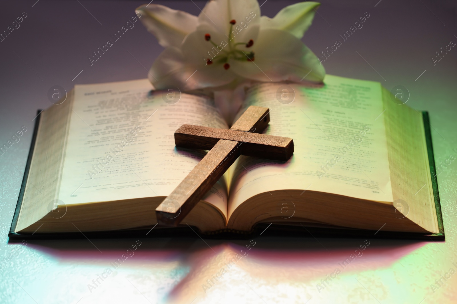 Photo of Wooden cross, Bible and lily flower on textured table in color lights, closeup. Religion of Christianity