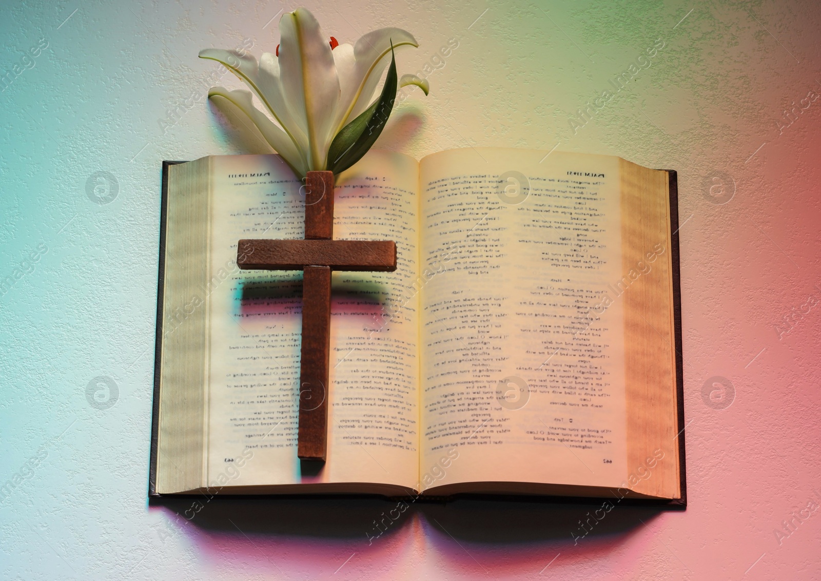 Photo of Wooden cross, Bible and lily flower on textured table in color lights, top view. Religion of Christianity