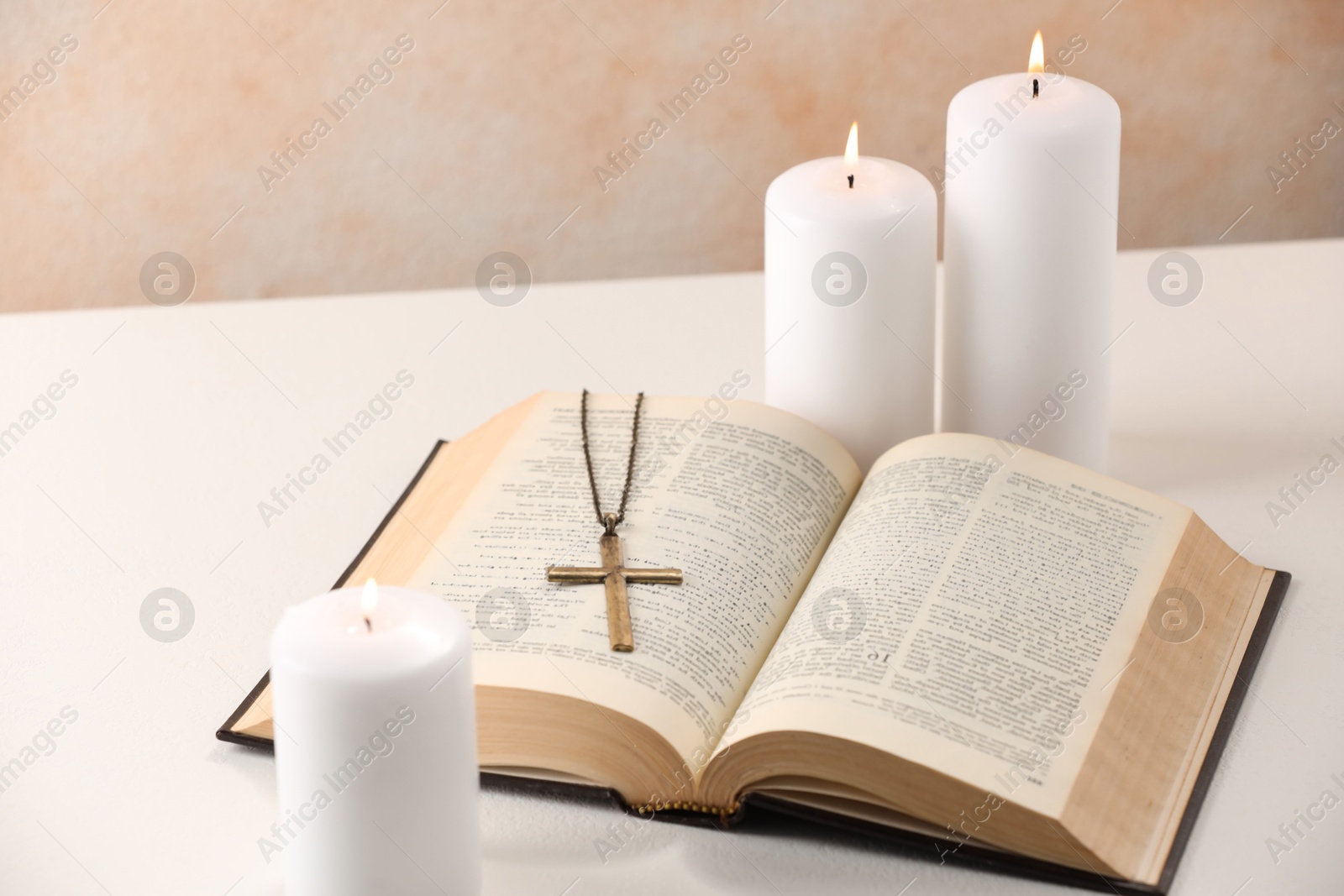 Photo of Cross with chain, burning candles and Bible on white table, closeup. Religion of Christianity