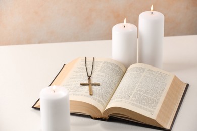 Photo of Cross with chain, burning candles and Bible on white table, closeup. Religion of Christianity