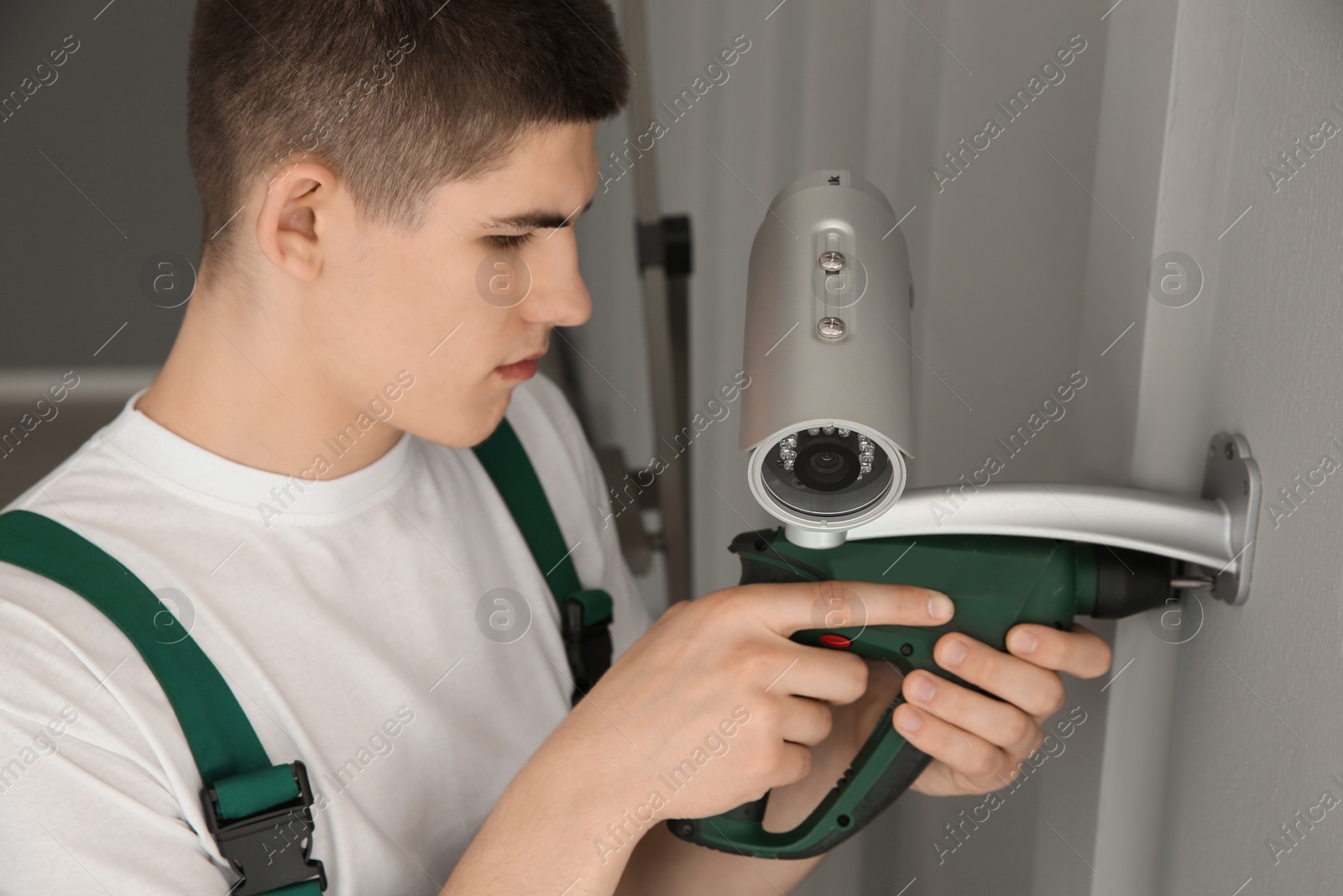 Photo of Technician with cordless electric screwdriver installing CCTV camera on wall indoors