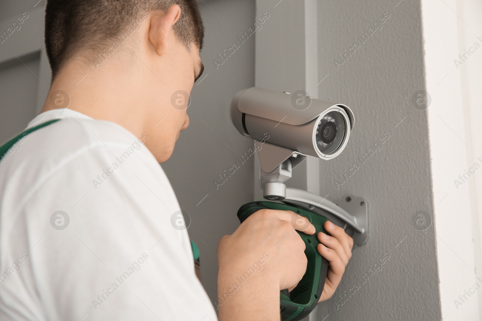 Photo of Technician with cordless electric screwdriver installing CCTV camera on wall indoors