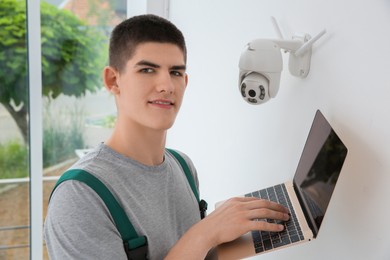 Technician with laptop installing CCTV camera on wall indoors