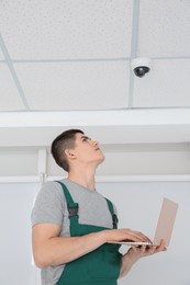 Technician with laptop installing CCTV camera on ceiling indoors, low angle view