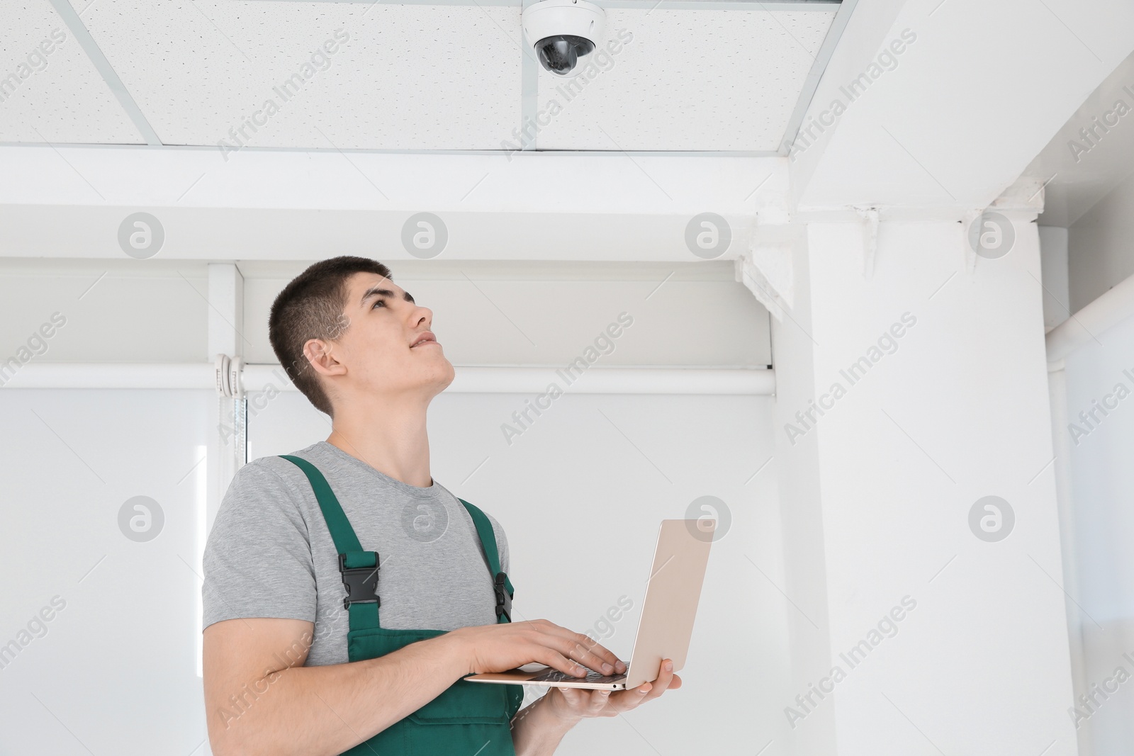 Photo of Technician with laptop installing CCTV camera on ceiling indoors