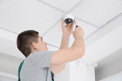 Technician with screwdriver installing CCTV camera on ceiling indoors, low angle view