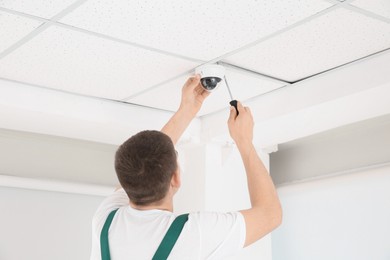 Technician with screwdriver installing CCTV camera on ceiling indoors, back view