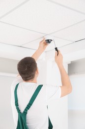 Photo of Technician with screwdriver installing CCTV camera on ceiling indoors, back view