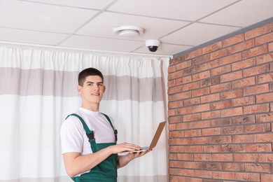 Technician with laptop installing CCTV camera on ceiling indoors