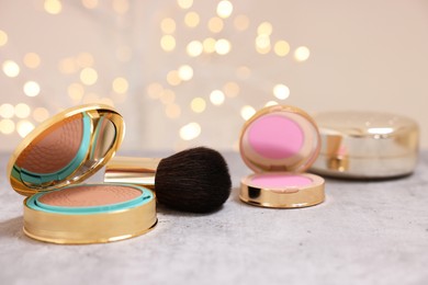 Photo of Face bronzer, blusher and other cosmetic products on grey textured table against blurred lights, closeup