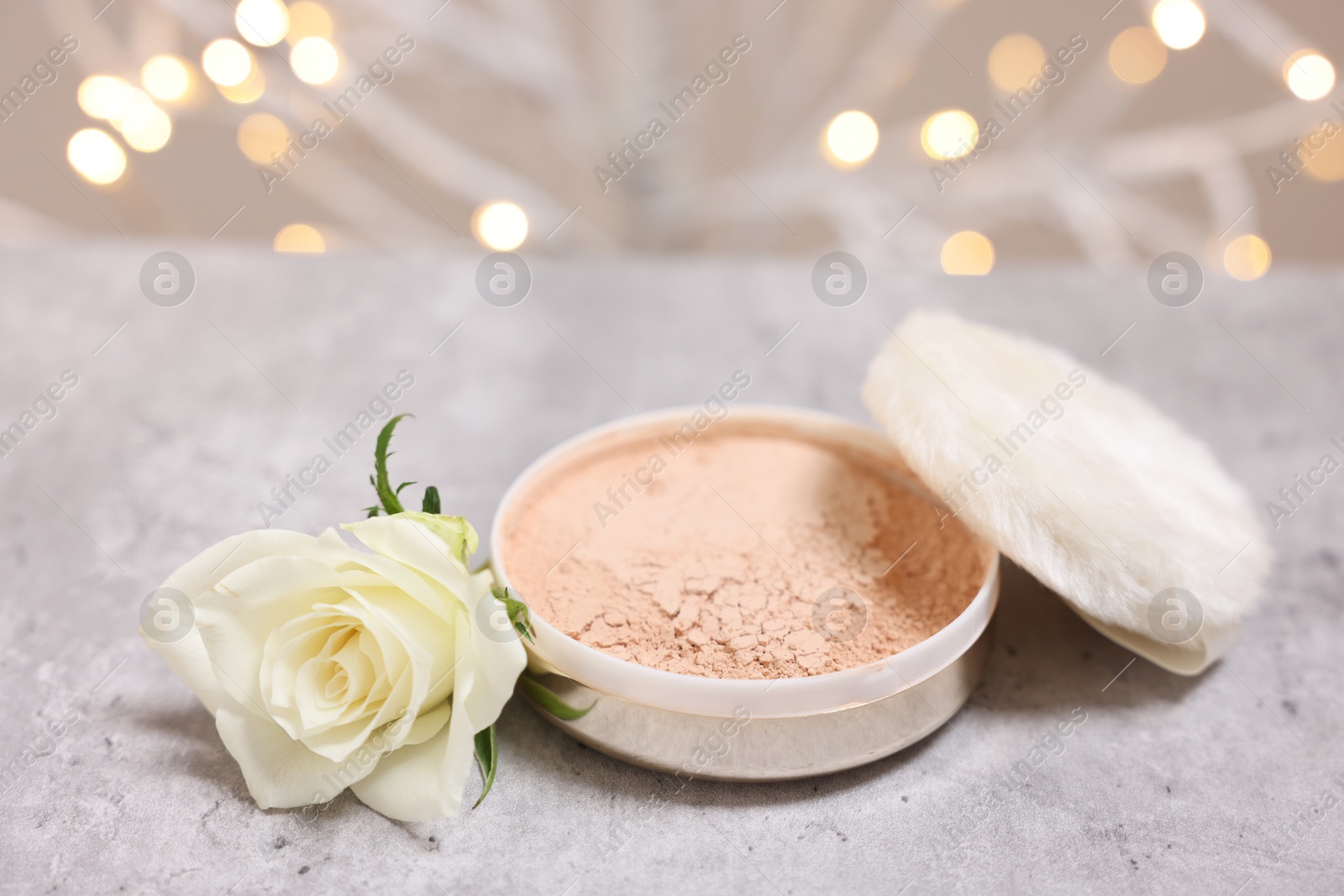 Photo of Face powder, puff applicator and rose flower on grey textured table against blurred lights, closeup