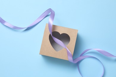 Beautiful violet ribbon and gift box with heart on light blue background, flat lay