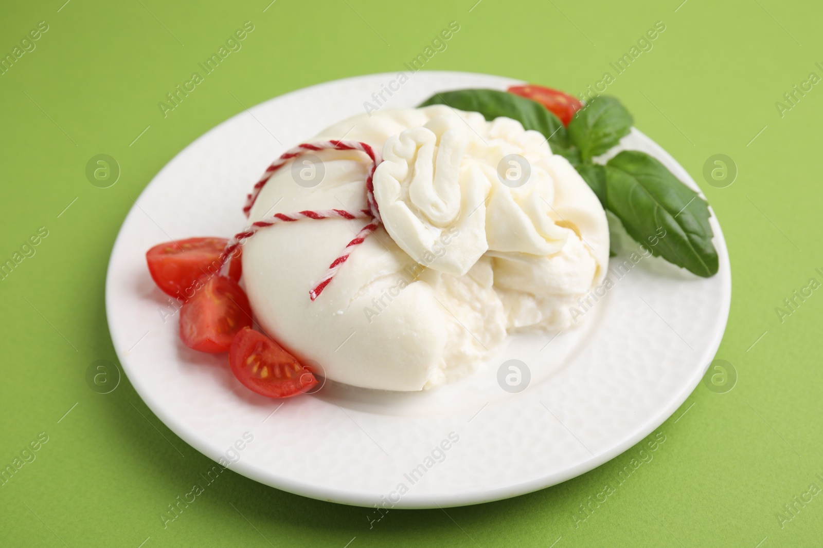 Photo of Delicious fresh burrata cheese with basil leaves and tomato on green table