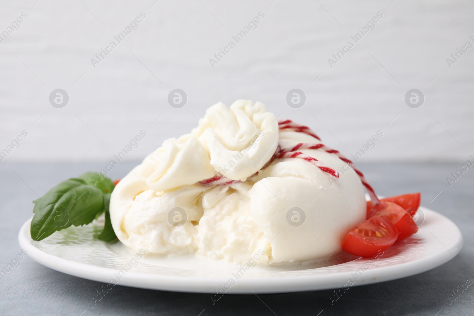 Photo of Delicious fresh burrata cheese with basil leaves and tomato on grey table, closeup