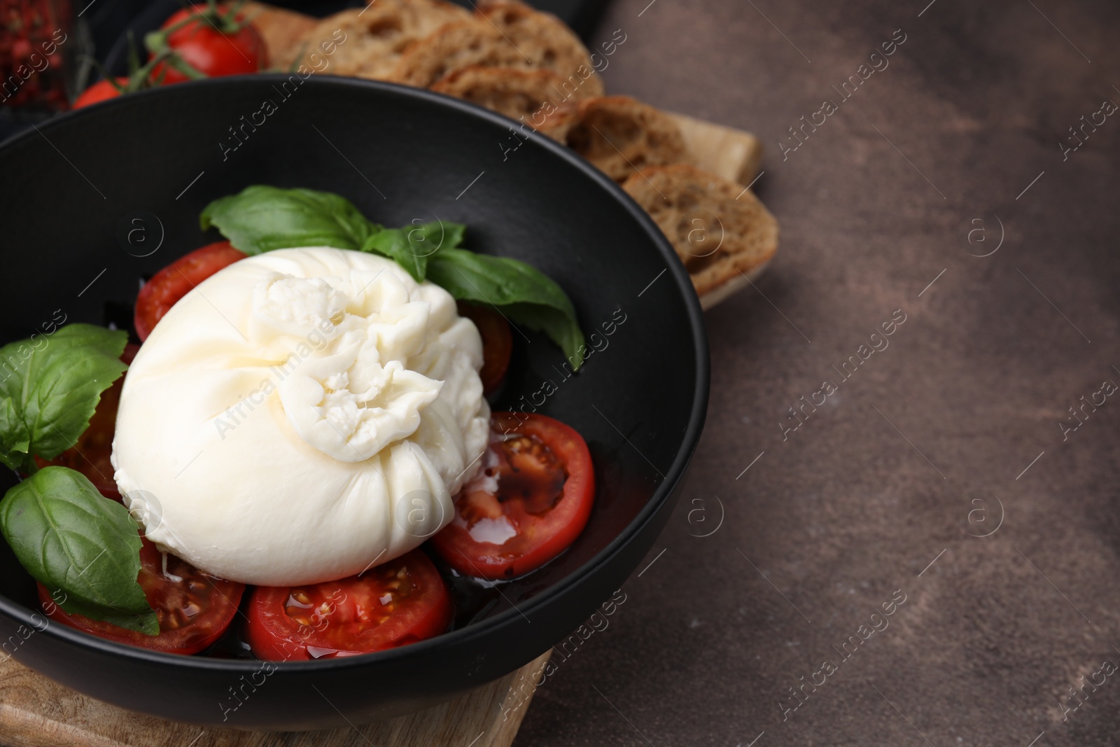 Photo of Delicious burrata cheese, tomatoes and basil in bowl on brown table, closeup. Space for text