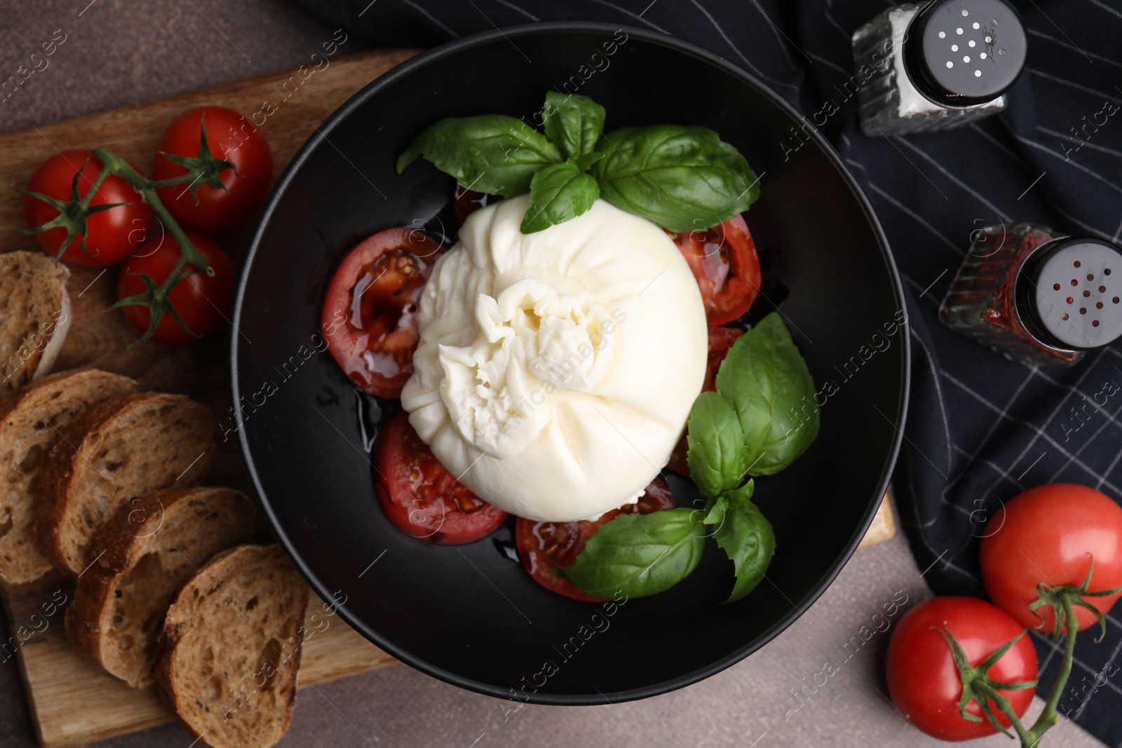 Photo of Delicious burrata cheese, tomatoes, basil and bread in bowl on brown table, flat lay