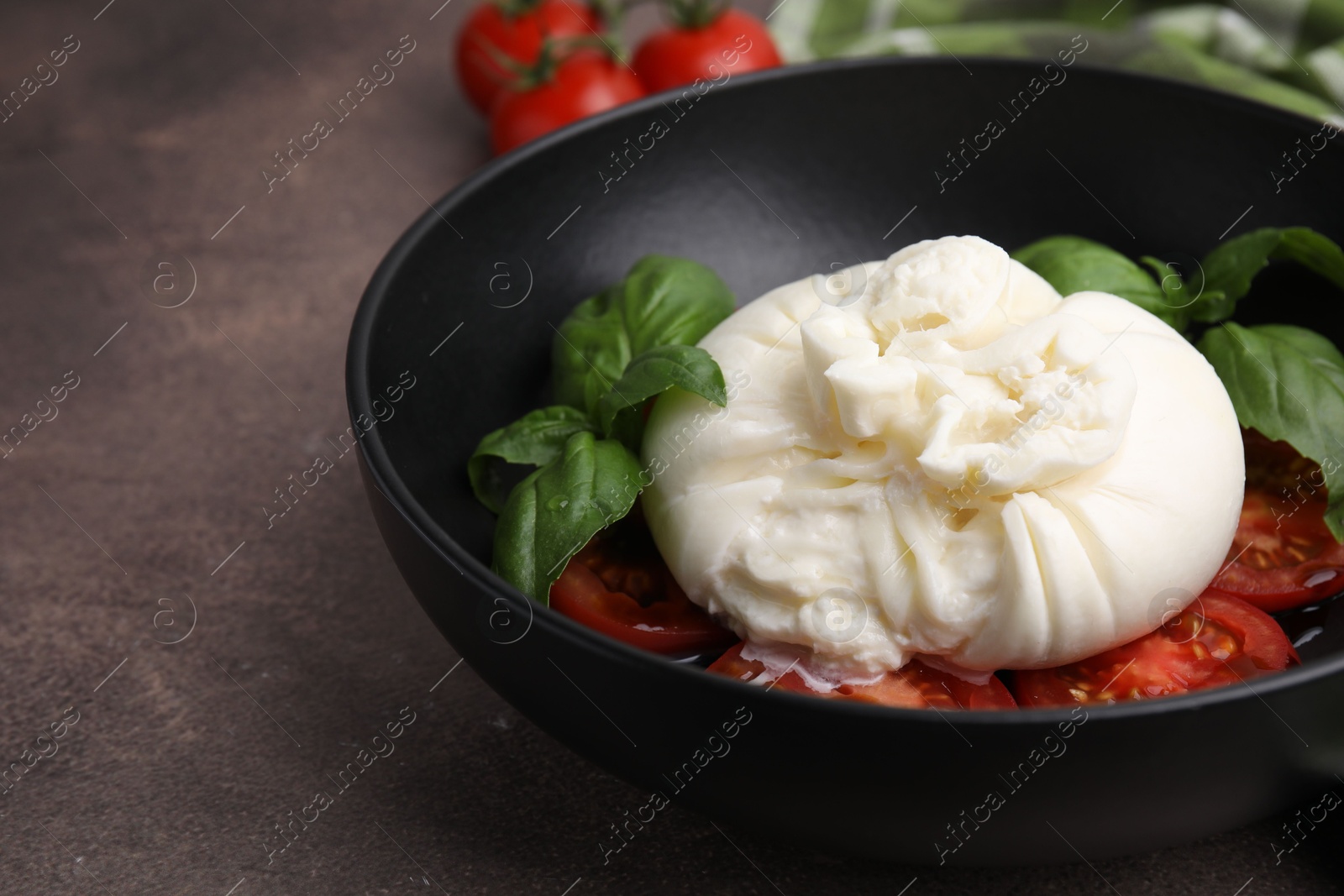 Photo of Delicious burrata cheese, tomatoes and basil in bowl on brown table, closeup