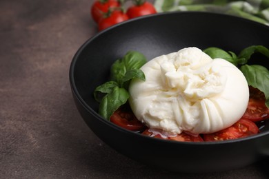 Photo of Delicious burrata cheese, tomatoes and basil in bowl on brown table, closeup