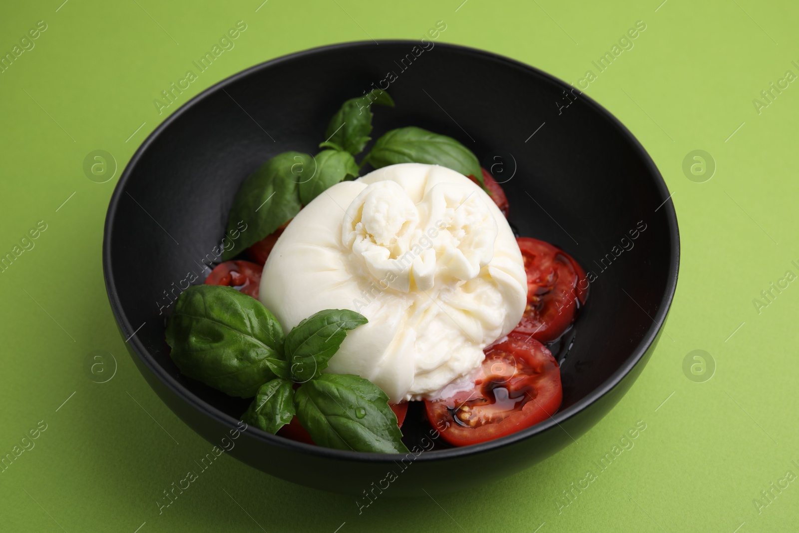 Photo of Delicious burrata cheese, tomatoes and basil in bowl on green table, closeup