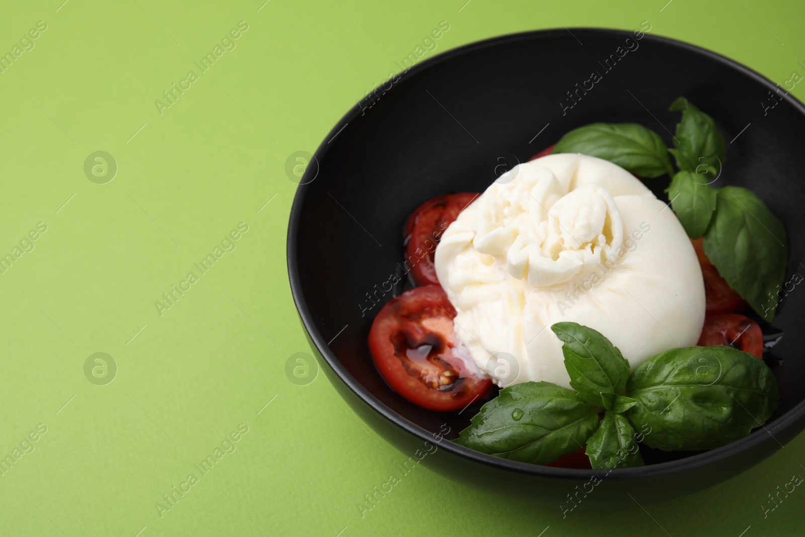 Photo of Delicious burrata cheese, tomatoes and basil in bowl on green table, closeup. Space for text