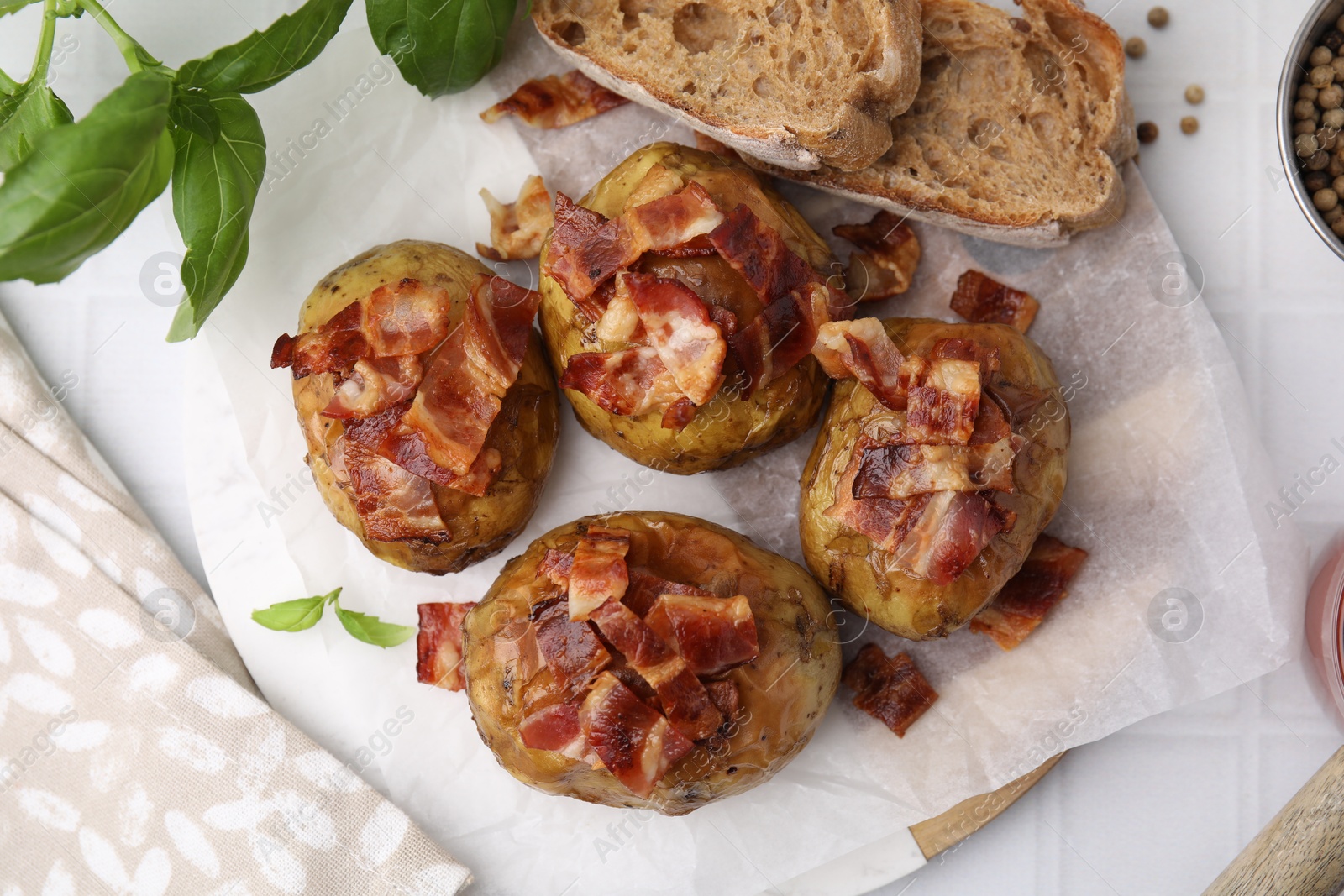 Photo of Delicious baked potatoes with bacon, bread and basil on white tiled table, flat lay