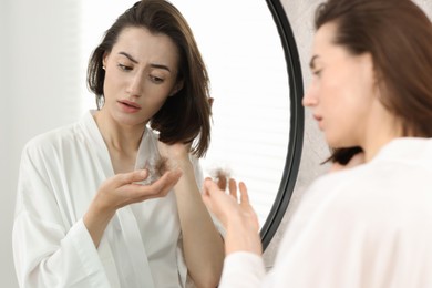 Photo of Sad woman holding clump of lost hair near mirror indoors. Alopecia problem