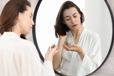 Sad woman holding clump of lost hair near mirror indoors. Alopecia problem