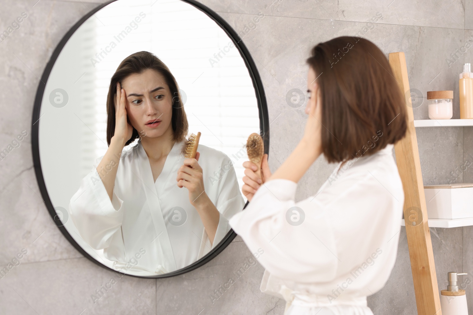 Photo of Stressed woman holding brush with lost hair near mirror at home. Alopecia problem