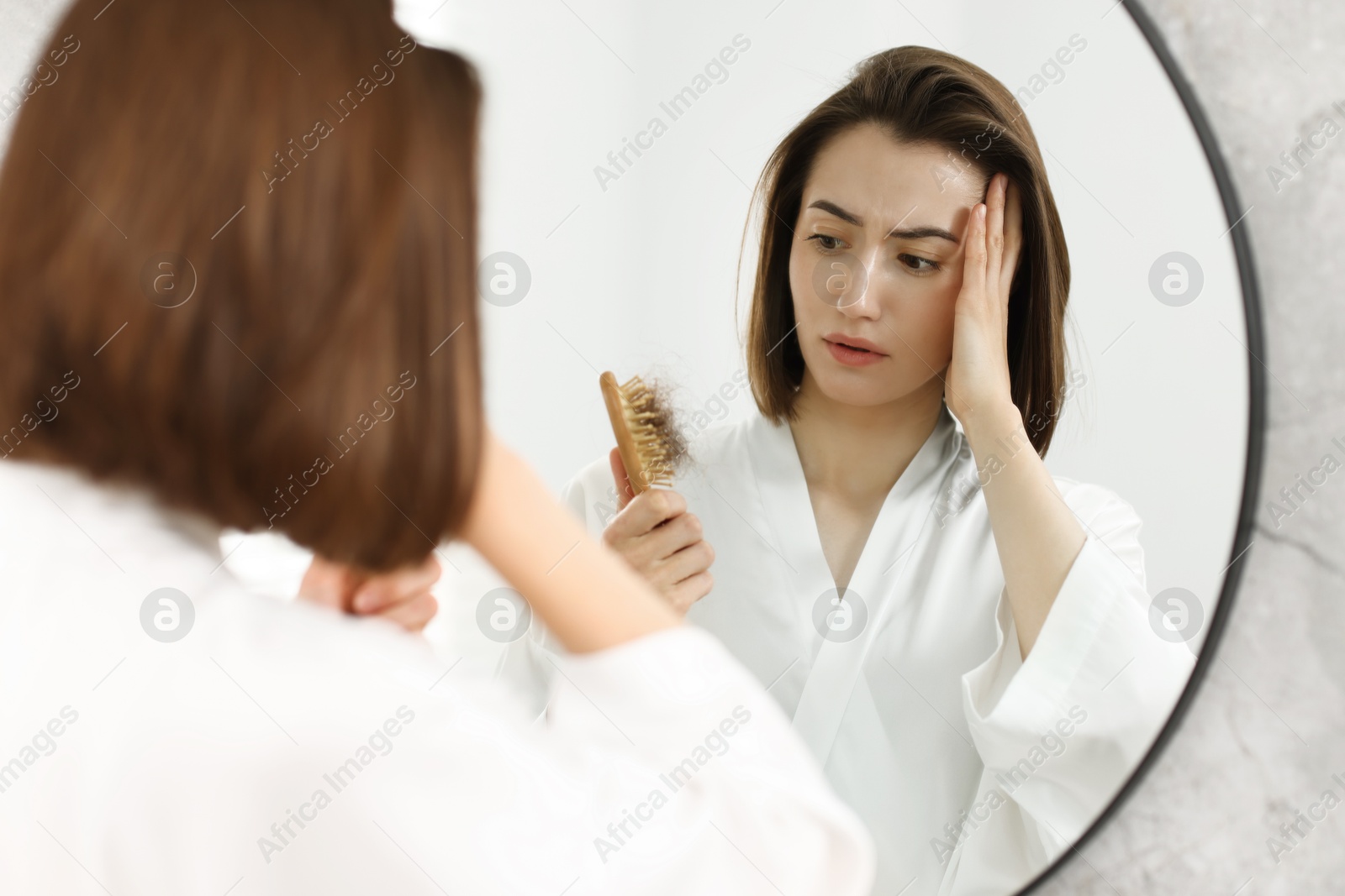 Photo of Stressed woman holding brush with lost hair near mirror at home. Alopecia problem