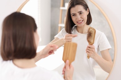 Photo of Emotional woman taking her lost hair from brush near mirror indoors. Alopecia problem