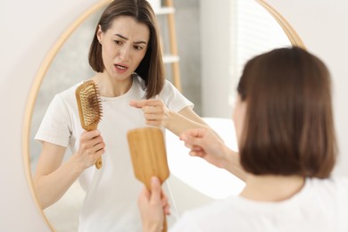 Emotional woman taking her lost hair from brush near mirror indoors. Alopecia problem