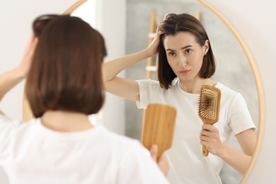 Sad woman holding brush with lost hair indoors. Alopecia problem