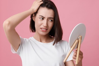 Sad woman with hair loss problem looking at mirror on pink background