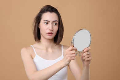 Sad woman with hair loss problem looking at mirror on light brown background