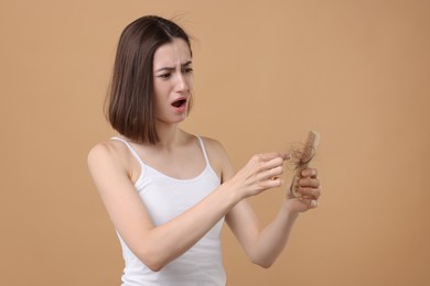 Emotional woman taking her lost hair from comb on light brown background. Alopecia problem