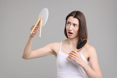 Emotional woman with hair loss problem looking at mirror on grey background