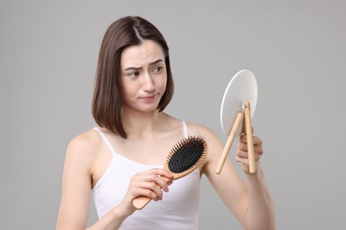 Sad woman with hair loss problem looking at mirror on grey background