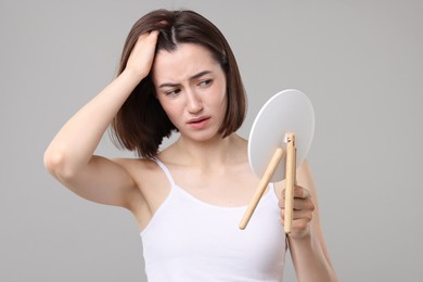 Sad woman with hair loss problem looking at mirror on grey background