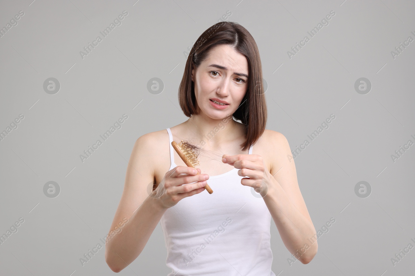 Photo of Emotional woman taking her lost hair from brush on grey background. Alopecia problem