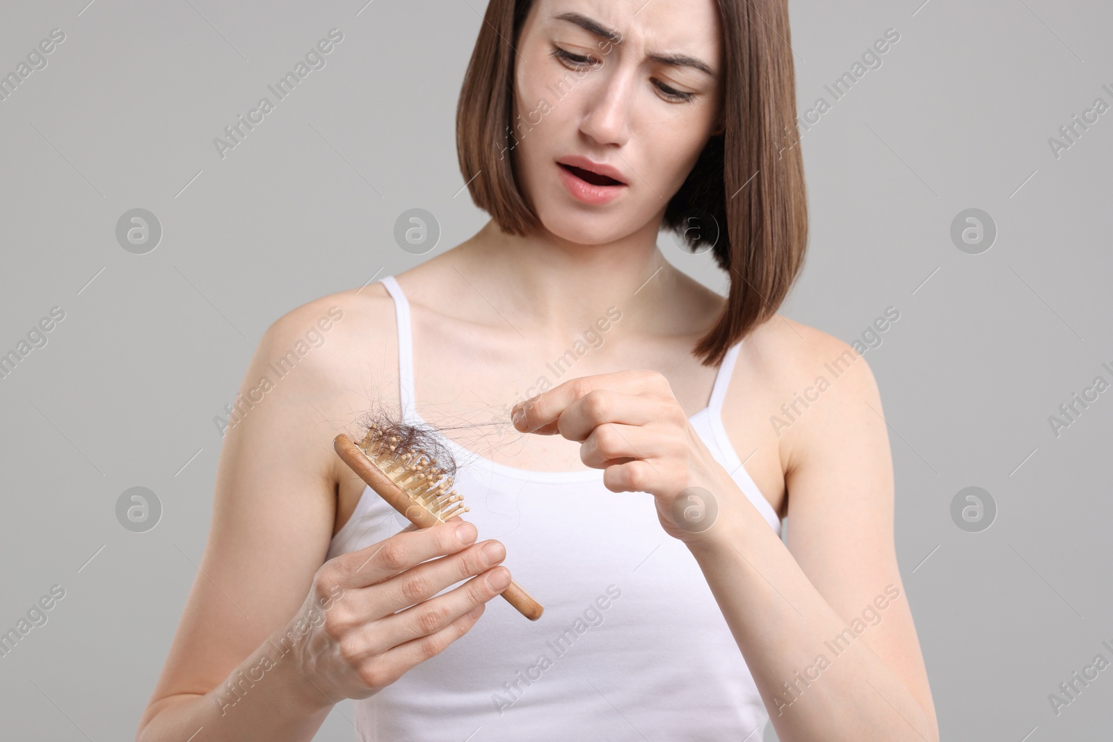 Photo of Stressed woman taking her lost hair from brush on grey background. Alopecia problem