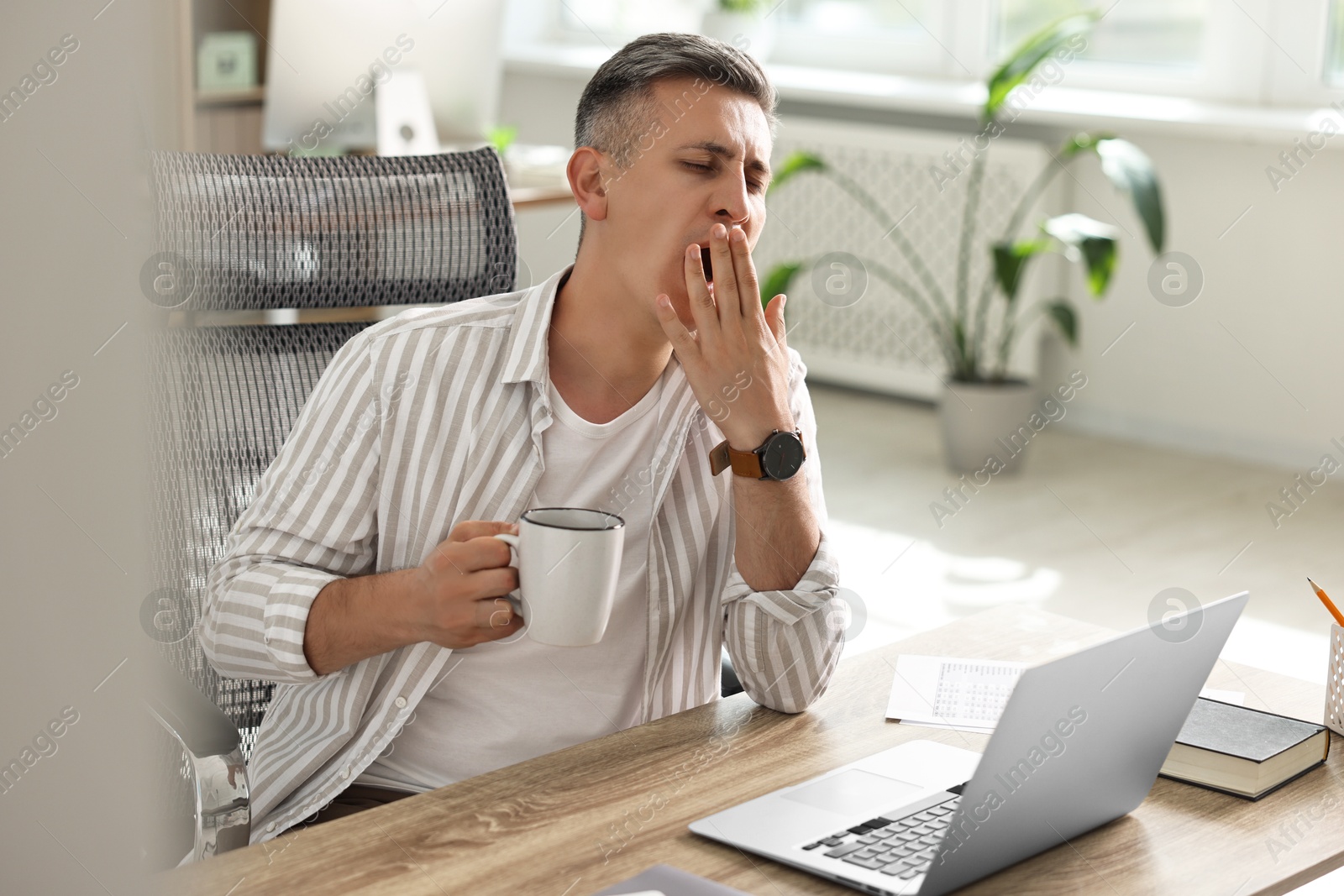 Photo of Man with cup of drink yawning at wooden table in office