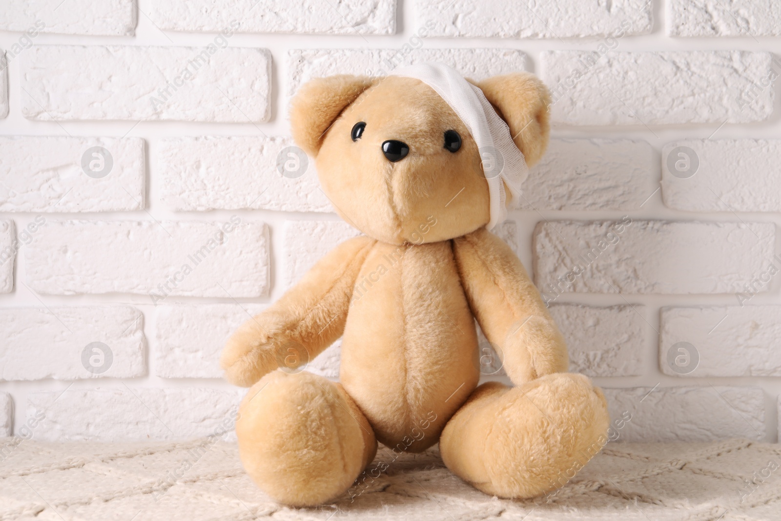Photo of Toy bear with bandage on soft surface near white brick wall