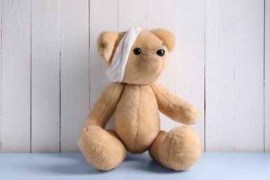 Photo of Toy bear with bandage on light blue table near white wooden wall