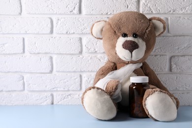 Toy bear with bandage and bottle of pills on light blue table near white brick wall, space for text