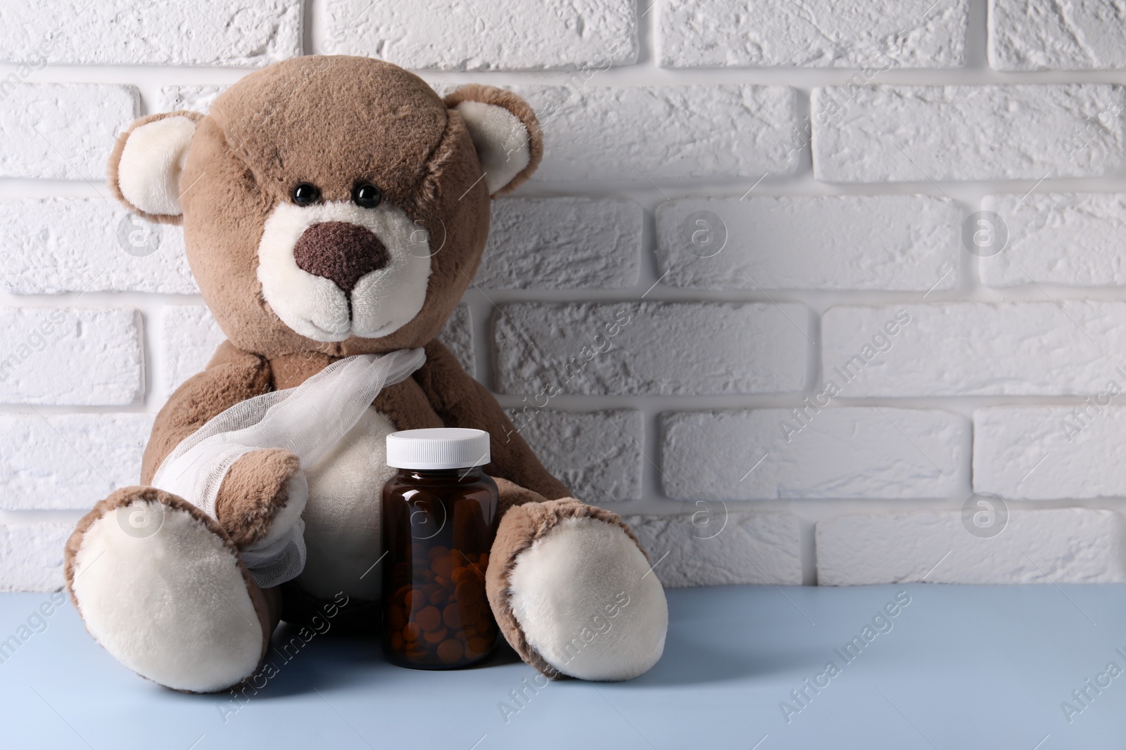Photo of Toy bear with bandage and bottle of pills on light blue table near white brick wall, space for text