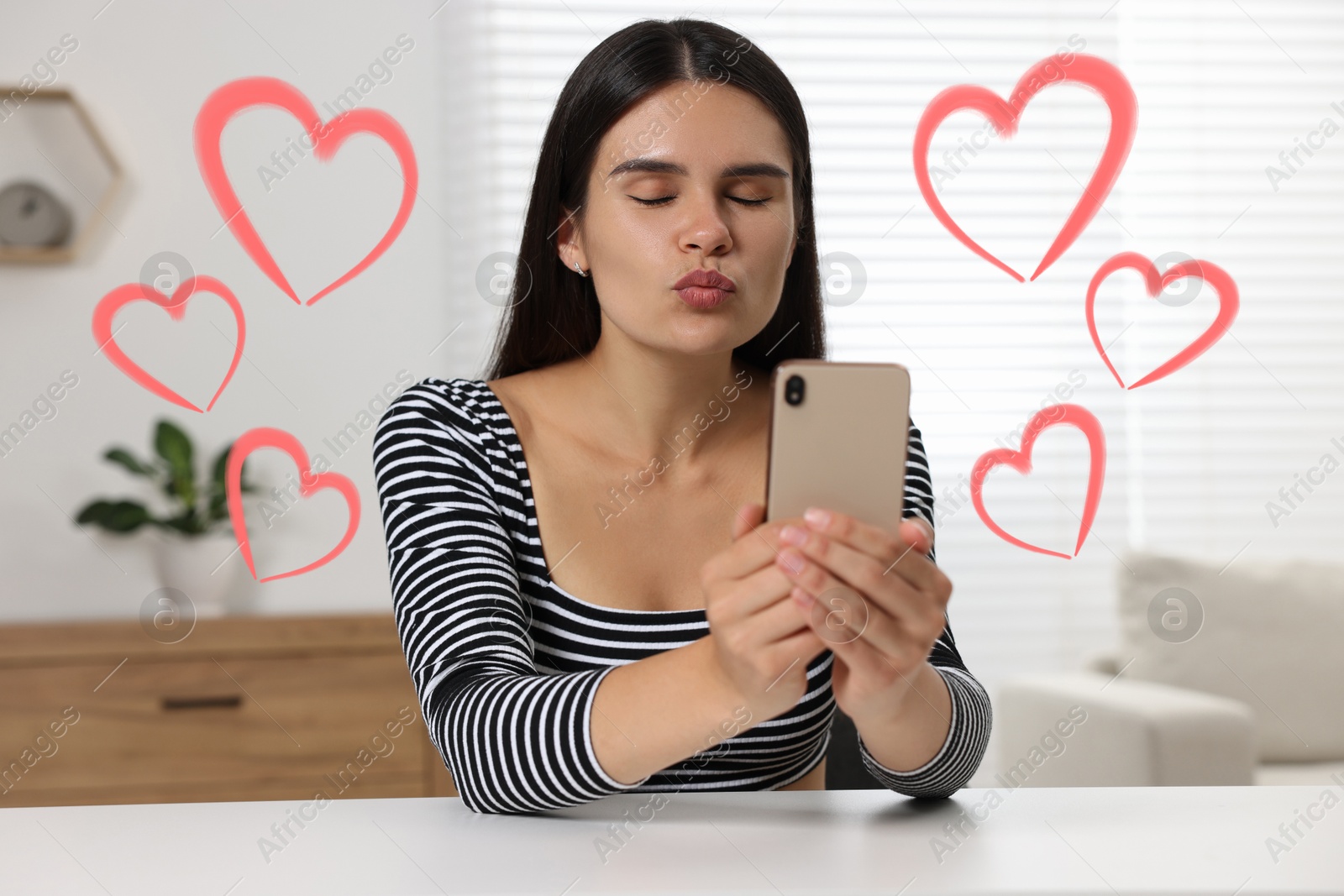 Image of Long distance relationship. Young woman sending air kiss to her loved one via video chat indoors. Red hearts around