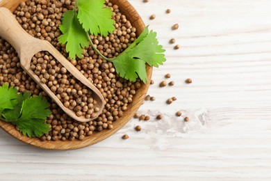 Dried coriander seeds with green leaves in bowl and scoop on wooden table, top view. Space for text