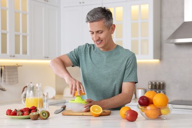 Photo of Smiling man squeezing fresh orange with juicer at white marble table in kitchen