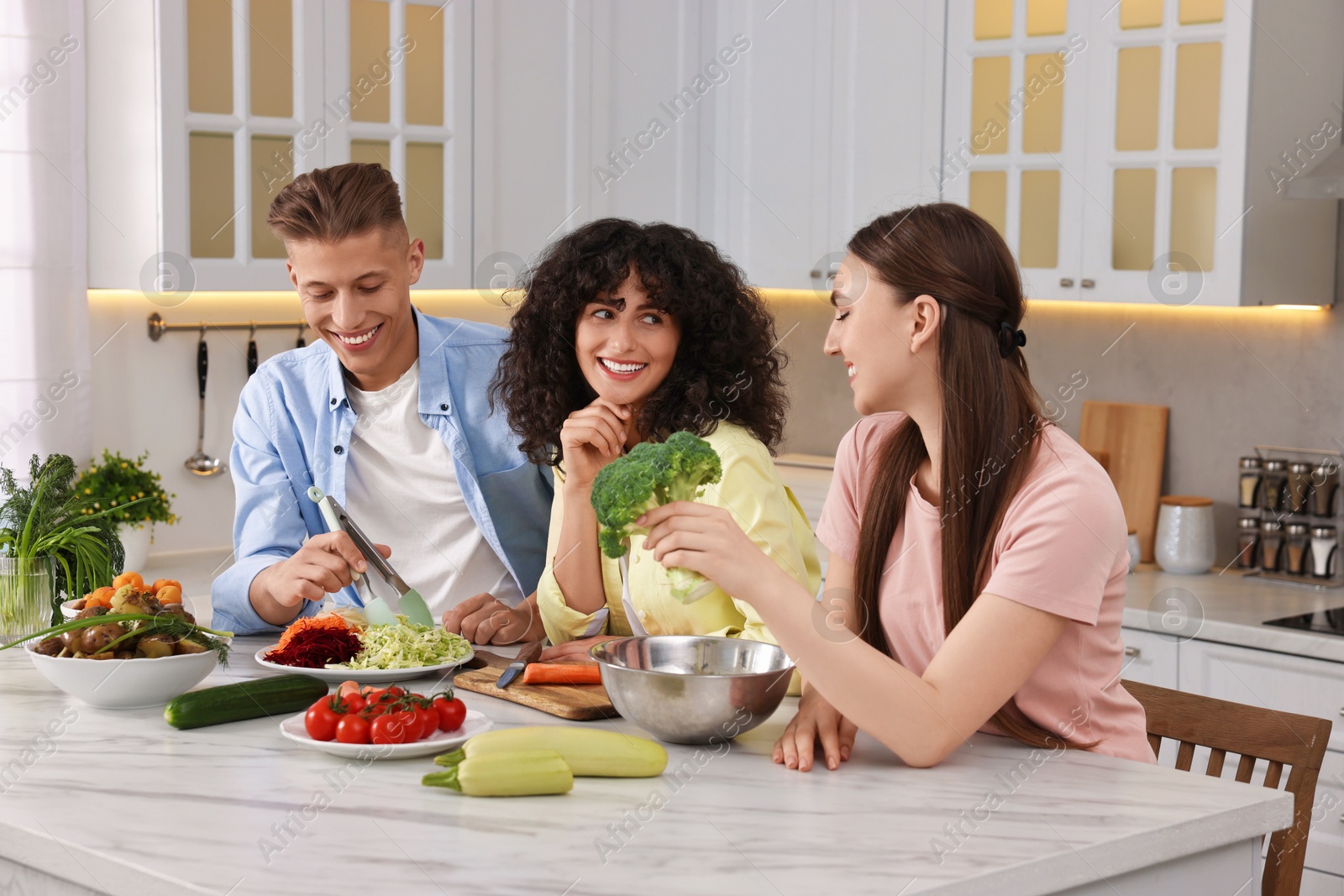 Photo of Friends cooking healthy vegetarian meal at white marble table in kitchen