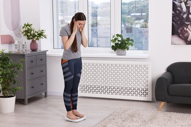 Photo of Worried woman standing on floor scale at home